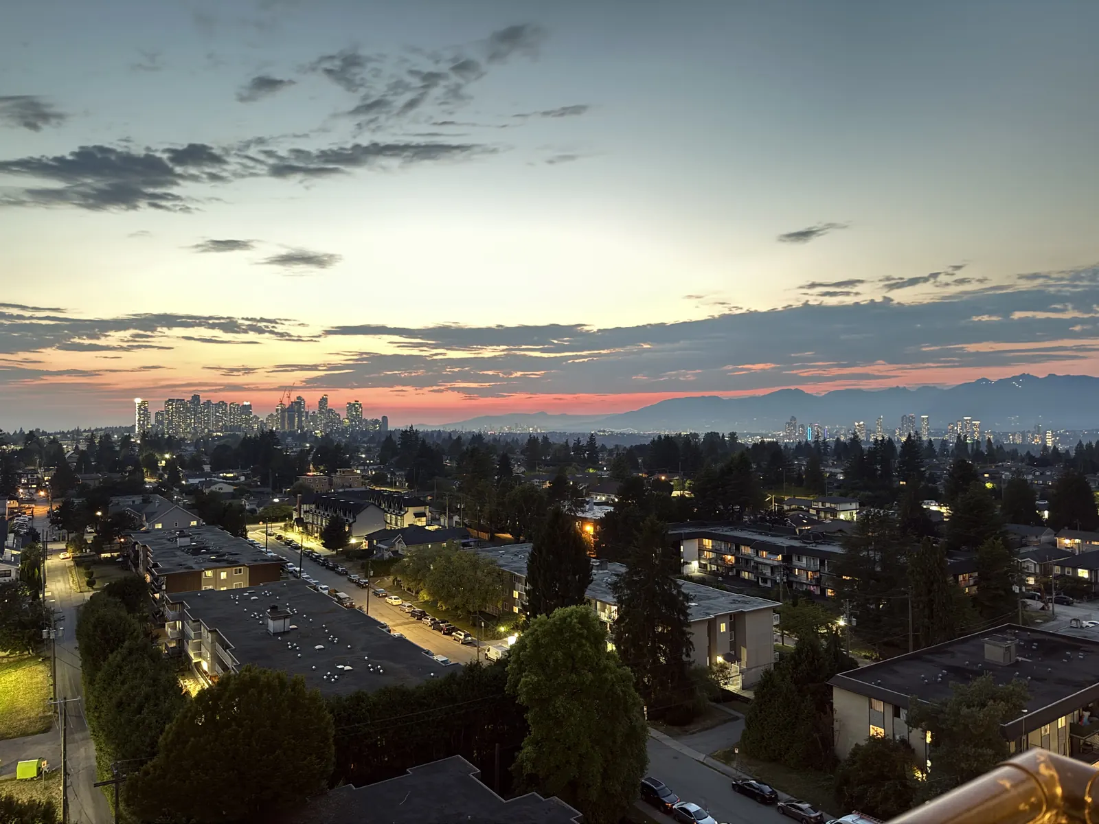 Dusk over Metro Vancouver from a high-rise, with streetlights, a distant skyline, and mountains on the horizon