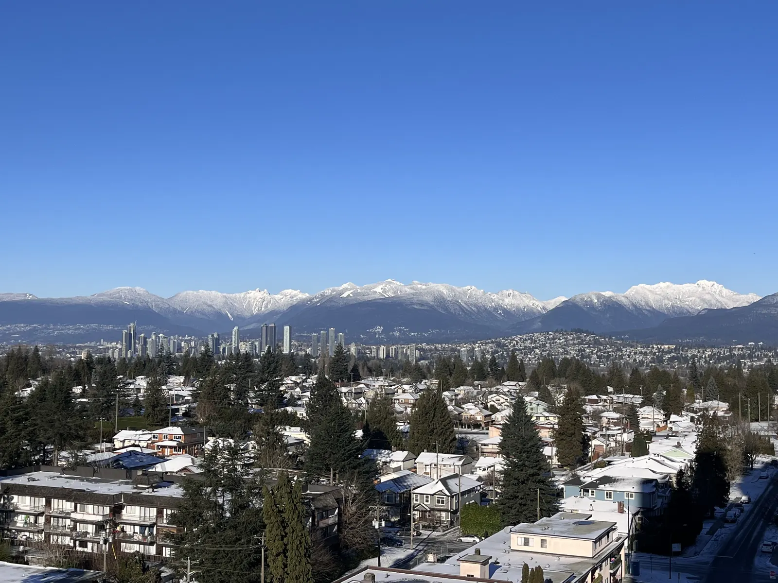 Winter view across a Metro Vancouver neighbourhood toward snow-capped North Shore mountains and a distant skyline